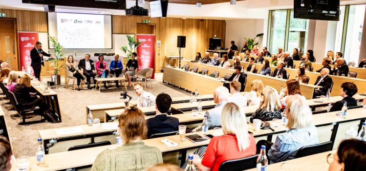 bright, modern lecture hall viewed from the back with presenter and seated panelists shown at the front and seated guests listening in